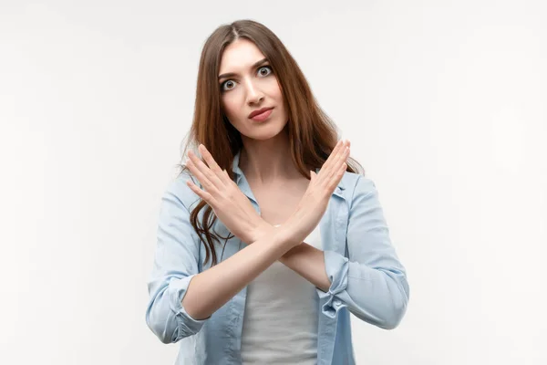 Image of serious woman 20s with long chestnut hair, making X sign with crossed hands, gesturing stop, warning of danger. No way, stop doing. Wearing casual clothes. Studio shot white background