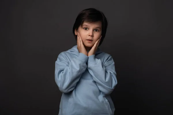 Image of shocked boy 10-12 years old, dressed in casual clothes expressing surprise on camera. Studio shot, gray background. Human emotions, facial expression concept