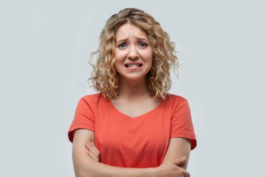 Utanmış, kıvırcık saçlı, sıradan tişörtlü, kameraya bakan sarışın kızın portresi. Stüdyo fotoğrafı, beyaz arka plan. İnsani duygular, yüz ifadesi kavramı