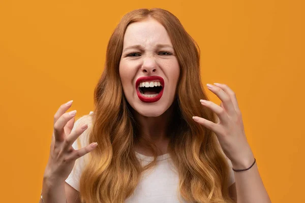 Angry young woman with wavy redhead, wearing white t shirt screaming on yellow studio background. Emotional face. Human emotions, facial expression concept.