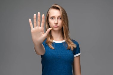 Mavi tişörtlü, elinde avuç içi ile dur işareti yapan ciddi sarışın bir kız resmi. Yüzünde uyarı ifadesi var. Stüdyo fotoğrafı, gri arka plan. İnsan duyguları yüz ifadesi kavramı