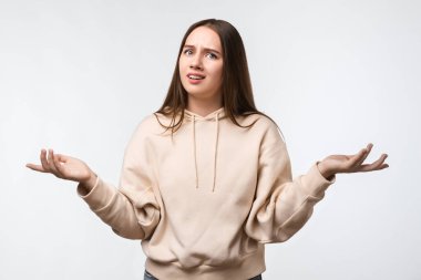 Uzun kestane rengi saçları olan, bej kapüşonlu şık giyinen, şüpheyle el ele tutuşan güvensiz genç bir kadının stüdyo fotoğrafı. Şüphe, belirsizlik ve karmaşa kavramı
