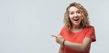 Kıvırcık saçlı, sıradan tişörtlü, gülümseyen ve işaret parmağıyla işaret eden mutlu genç bir kadın görüntüsü reklam için uygun bir yer. Stüdyo çekimi, beyaz arkaplan, izole edilmiş