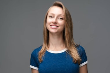 Studio shot of pretty female with blonde straight hair, broad smile, wears blue t shirt, tilts head and looks with joy. Human emotions, facial expression concept. clipart