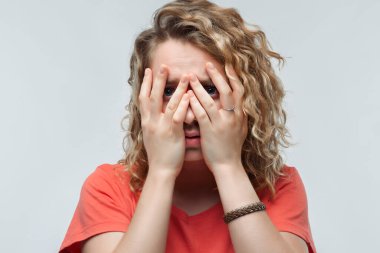 Sıradan tişörtlü, korkmuş, kıvırcık saçlı, elleriyle gözlerini kapatan genç bir kadının portresi. İnsani duygular, yüz ifadesi kavramı. Stüdyo çekimi, beyaz arkaplan, izole edilmiş