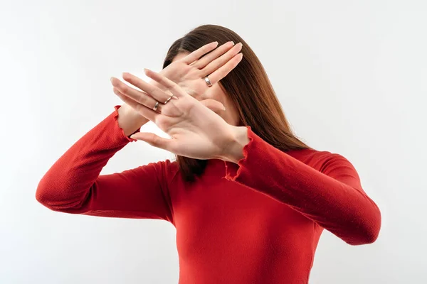 Image of serious woman with long chestnut hair in casual red sweater gestures actively hands at camera, stop gesture with hands. No way, stop doing. Studio shot, white background. Human emotions concept