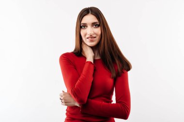 Uzun kestane rengi, sıradan kırmızı kazaklı ve kameraya bakan utangaç genç bir kadının portresi. Stüdyo fotoğrafı, beyaz arka plan. İnsani duygular, yüz ifadesi kavramı