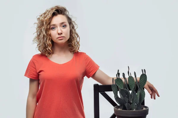 Young beautiful woman with curly hairstyle in casual t shirt smiles and holds cactus in her hands, potted green plant. Studio shot, white background. Home plants care concept