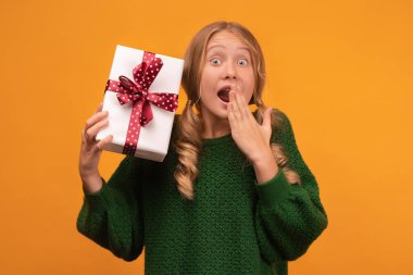 12-14 yaşlarında, sıcak yeşil kazaklı, elinde kırmızı fiyonklu hediye kutusu olan çekici bir sarışın kız resmi. Stüdyo fotoğrafı, sarı arka plan, izole edilmiş. Yeni yıl kadınlarının doğum günü konsepti