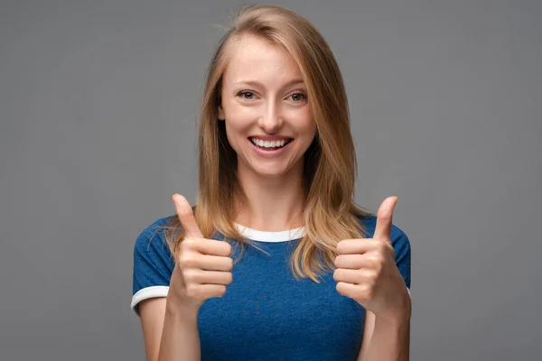 Cheerful girl with blonde straight hair looking at the camera with happy expression, showing thumbs-up with both hands, excellent job. Human emotions, facial expression concept.