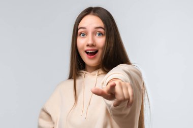 Uzun kestane rengi saçları olan, günlük kıyafetler giymiş, kameraya bakan, gülümseyen ve seni işaret eden güzel bir kız.