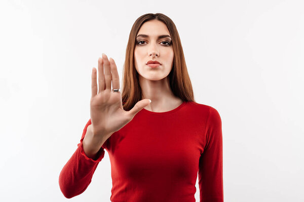 Image of serious woman with long chestnut hair in casual red sweater doing stop sign with palm of the hand. Warning expression on the face. Studio shot, white background. Human emotions concept