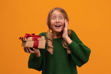 12-14 yaşlarında, sıcak yeşil süveter giyen, gülümseyen ve kırmızı kurdeleli hediye kutusunu tutan çekici bir sarışın kız resmi. Stüdyo fotoğrafı, sarı arka plan. Yeni Yıl Kadınların Doğum Günü Tatil Konsepti