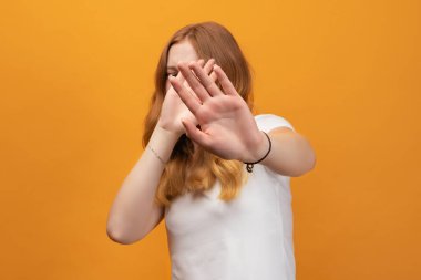 Dalgalı kızıl saçlı, beyaz tişörtlü, elleri ve gözleri kapalı güzel bir kız. Utanç verici ve olumsuz bir kavram, sarı arka planda izole edilmiş.