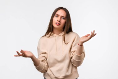 Uzun kestane rengi saçları olan, bej kapüşonlu şık giyinen, şüpheyle el ele tutuşan güvensiz genç bir kadının stüdyo fotoğrafı. Şüphe, belirsizlik ve karmaşa kavramı