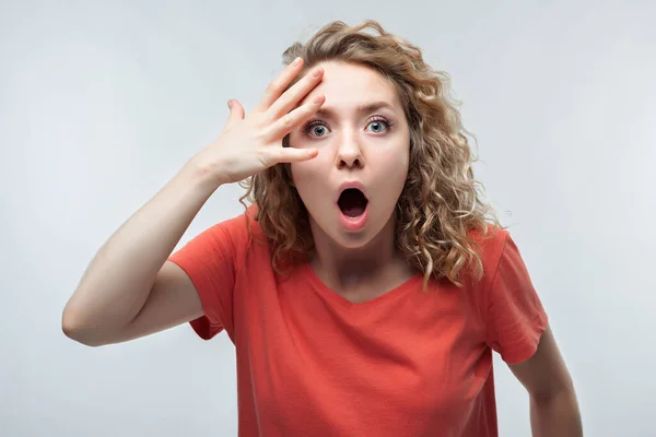 Şok olmuş, kıvırcık saçlı, sıradan tişörtlü, kameraya sürpriz yapan genç bir kadın. İnsani duygular, yüz ifadesi kavramı. Stüdyo çekimi, beyaz arkaplan