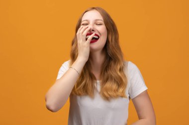 Dalgalı kızıl saçlı, beyaz tişörtlü, beyaz dişlerini gösteren neşeli genç bir kadın. Stüdyonun sarı arka planında pozlar veriyor. İfade ve zevkten zevk alıyor. Yüz ifadeleri