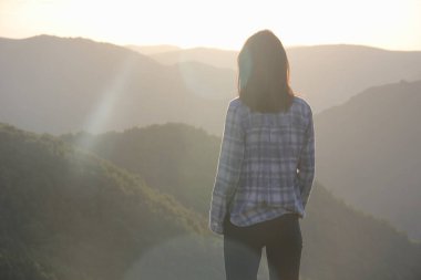 Altın sarısı günbatımı ışığı içindeki genç kadın uzaklardaki dağların arka planına bakıyor..