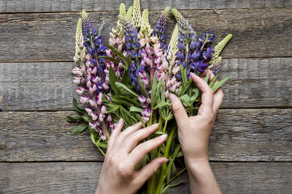 Women\'s hands are collected in a bouquet of lupine flowers on a wooden background.
