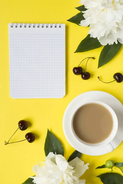 Notepad for text white peony flowers cherry berries Cup of coffee on yellow bright background