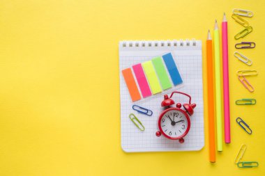 Defter, kalem ve renkli ataç, sarı zemin üzerine çalar saat.