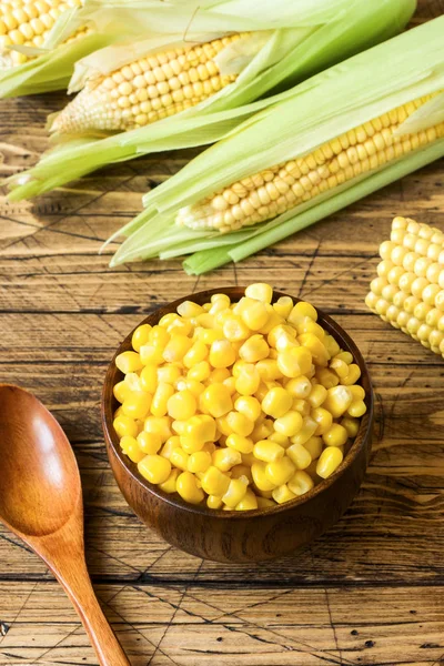 Canned corn in a wooden plate and cob of fresh corn on a rustic wooden background.