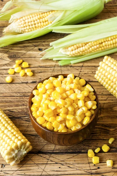 Canned corn in a wooden plate and cob of fresh corn on a rustic wooden background.