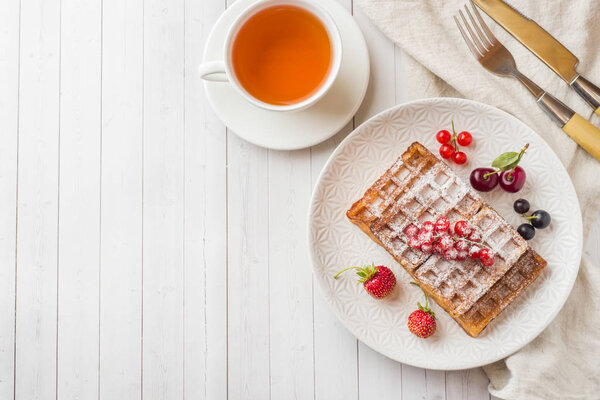 Homemade waffles with summer berries on a plate. A Cup of tea on a light table. Selective focus.