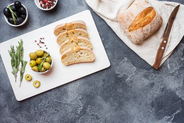 올리브와 로즈마리를 곁들인 이탈리아 식 시아바타 빵을 도마 위에 올렸습니다. 어두운 콘크리트 배경입니다. 복사 공간. — 스톡 사진