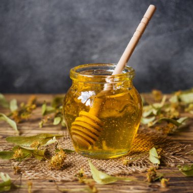 Ihlamur çiçeklerinden bir kavanoz sıvı bal ve koyu renkli bir arka plan üzerinde bal olan bir çubuk. Kopyalama alanı