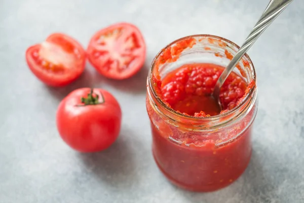 Tomato paste canned in a glass jar and fresh tomatoes on a concrete background. Fermentation of products. — Stock Photo, Image