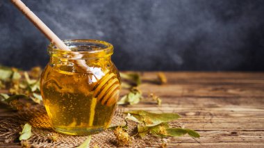 Ihlamur çiçeklerinden bir kavanoz sıvı bal ve koyu renkli bir arka plan üzerinde bal olan bir çubuk. Kopyalama alanı