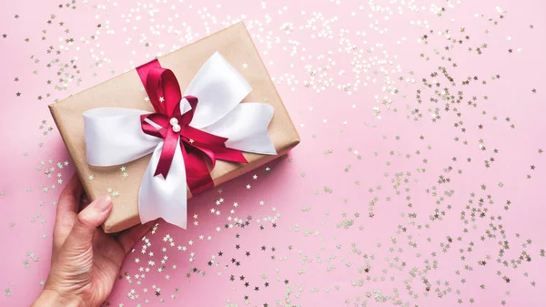 Concept of giving a gift. A box with a bow is handed by a womans hand. Pink background with Copy space. — Stock Photo, Image