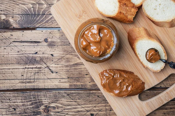 Gekochte Kondensmilch mit einem Glasgefäß auf einem hölzernen Hintergrund. Süße Paste auf Brotscheiben zum Frühstück und zum Dessert. Kopierraum — Stockfoto