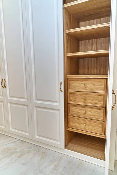 Opened white wardrobe with wooden drawers and shelves. Wooden filling of wardrobe. Wooden wardrobe with white lacquered cabinet doors