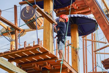 Miğferli ve tırmanma ekipmanlarıyla gökyüzüne karşı tahta bir platformda duran, kablo parkında vakit geçiren bir çocuk.