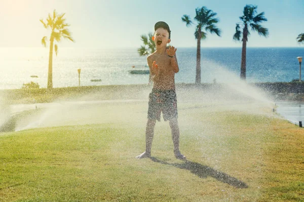 キャップとシャツのフルボディのシャツの少年は リゾートの晴れた夏の日に芝生の上でスプリンクラーで遊んでいる間叫んでいます — ストック写真