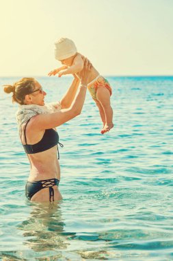 Mayo giymiş neşeli bir kadın bebeğiyle oynuyor ve tatil köyündeki deniz dalgalarının arka planına gülüyor.