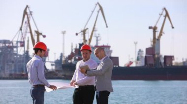 Bir grup mühendis, limanda vinçlerin arka planına karşı bir inşaat planını tartışıyorlar..