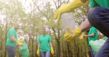 Bir gönüllü plastik bir şişeyi çevirir ve çöp torbasına atar. Parkta birlikte temizlik yapan diğer gönüllülerin arka planına karşı..