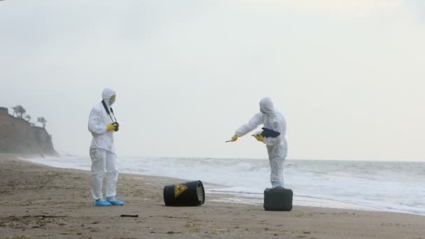 Two Men White Protective Suits Inspecting Seashore Black Barrel Sign — Stock Video