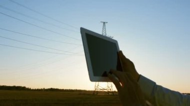 Özel kıyafetli bir güç mühendisi elinde bir tablet ile yüksek voltajlı bir elektrik hattının arka planında, durumunu inceliyor..