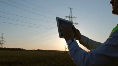 Özel kıyafetli bir işçi elektronik tablete not alır tableti indirir ve yüksek voltajlı elektrik hattının metal kulesine bakar..