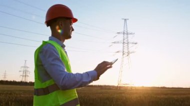 Elektronik tabletli bir işçi yüksek voltajlı elektrik hattının önünde yürüyor ve tablet üzerindeki direklerin fotoğraflarını çekiyor..