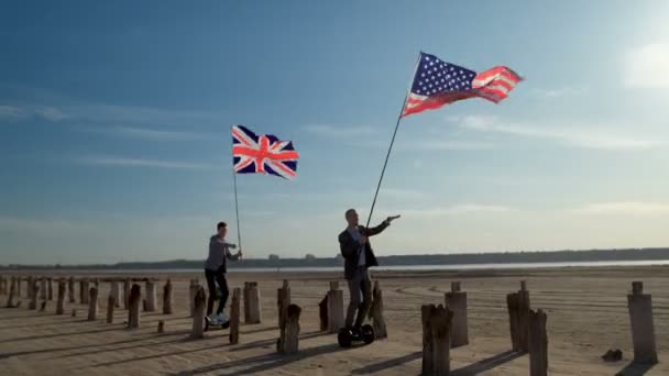 两个拿着美国和英国国旗的商人兴高采烈地骑着陀螺滑板车沿着海湾岸走着 — 图库视频影像