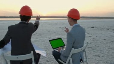 İş kıyafetleri ve turuncu kasklı iki adam ABD bayrağının altındaki boş bir alanda sandalyelere oturuyor. İnşaat planı tartışılıyor. Gayrimenkule yatırım.