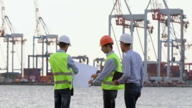 Baş mühendis tabletteki çalışma planını bir mühendis ve işçiye gösteriyor. Koruyucu miğferli adamlar limandaki bir posterde çizim yaparlar..