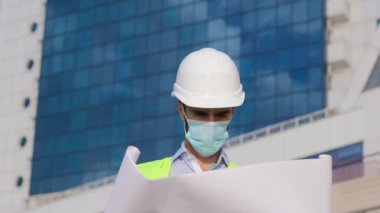 Yüzünde maske ve beyaz koruyucu kasklı bir adam ellerinde bir poster tutuyor ve mavi pencereli bir binanın arka planına bakıyor..
