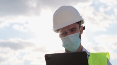 Yüzüne maske takan bir mühendis tabletle çalışır, arka planda alt köşeden vurulur. Bir depo çalışanı lojistiği tablet kullanarak kontrol eder..