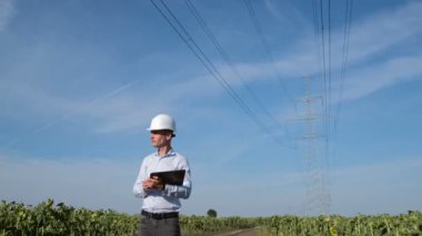 Beyaz korumalı miğferli, elektronik tabletli bir adam, yüksek voltajlı elektrik hattının önündeki ayçiçeği tarlalarının arasında yol boyunca yürüyor..